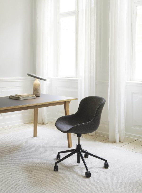 Et minimalistisk kontor med en mørk polstret stol ved et træskrivebord. Værelset, en udførelsesform af TomorrowsDesign, har hvide vægge, høje draperede vinduer og et tæppe med lyst område. En enkelt bog og en lille skulptur sidder på skrivebordet.
