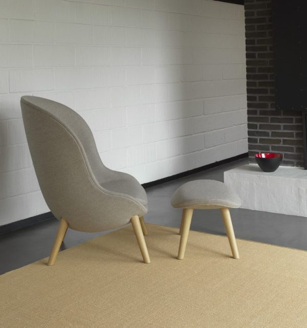 En moderne grå wingback stol med træben og en matchende skammel på et beige tæppe, placeret i et rum med hvide murstensvægge og en sort murstenssøjle. Et hvidt bord med en rød skål er i baggrunden. Denne opsætning viser enkle, men elegante designmøbler.
