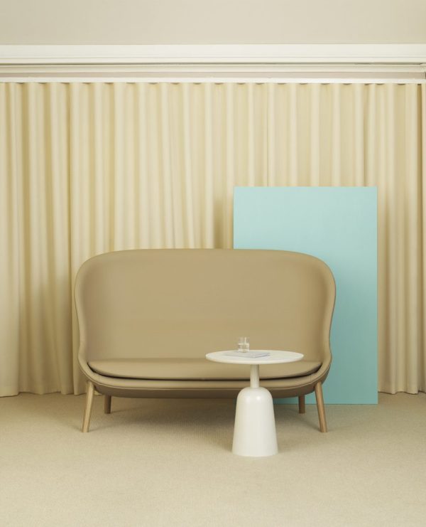 Et minimalistisk værelse med en beige sofa mod et cremet gardinbagtæppe, sammen med et lille NORMANN COPENHAGEN - Turn Bord. et lyseblåt panel er placeret bag sofaen, delvist synligt. den overordnede stemning er rolig og moderne.