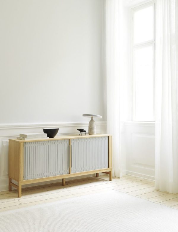 Et minimalistisk stuehjørne med en træskænk med lameldøre mod en hvid væg, nær et højt vindue draperet med gennemsigtige gardiner. Rummet har et hvidt gulv og et enkelt tæppe med et par elegante vaser oven på skænken, der viser moderne interiørdesign.