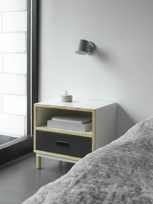 Et minimalistisk soveværelseshjørne med en grå seng, et NORMANN COPENHAGEN - Kabino Natbord natbord med en skuffe og en hylde med bøger, en hvid kop på toppen og en moderne grå væglampe.