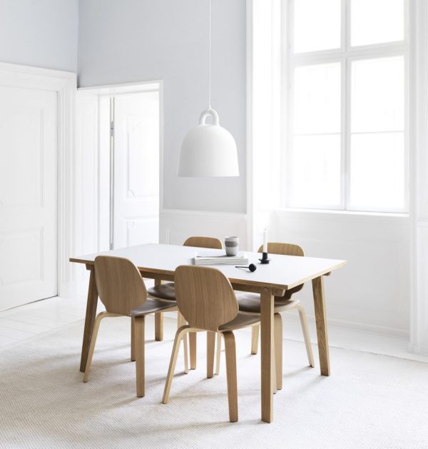En minimalistisk spisestue med en NORMANN COPENHAGEN - Bell Lampe Ø42 hængende over et lyst træbord med fire matchende stole, sat på et hvidt vævet tæppe. rummet har store vinduer og lysegrå vægge.