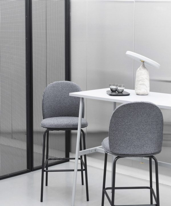 Et moderne minimalistisk kontorlokale med en grå polstret stol ved et hvidt bord, et NORMANN COPENHAGEN - Eddy Bordlampe og tesæt på toppen, sat op mod en mønstret sort/hvid skillevæg og persienner.