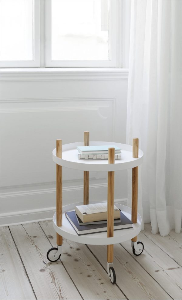 En lille to-lags hvid og træ vogn på hjul, designet af TomorrowsDesign, placeret ved et vindue med et gennemsigtigt gardin, rummer bøger og en bakke med dominobrikker på et lyst trægulv.