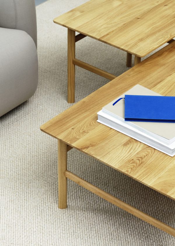 Et hyggeligt stuehjørne med to designmøbler-borde i træ. En blå hardcover bog og hvid notesbog hviler på det nederste bord, sat mod et blødt beige tæppe og en del af en grå sofa.