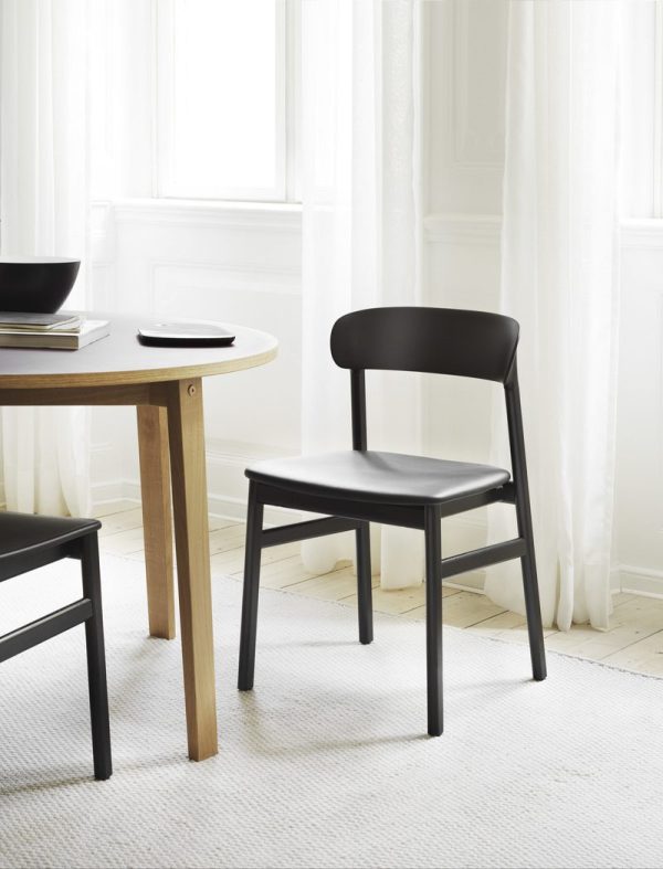 Et minimalistisk interiørdesign med en sort stol ved siden af et rundt træbord med bøger, sat på baggrund af store hvide gardiner og et lyst værelse.