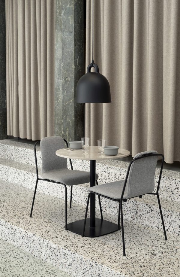 En moderne spiseplads med en NORMANN COPENHAGEN - Bell Lampe Ø42 med to grå polstrede stole, sat på baggrund af marmorvægge og tunge beige gardiner, under en stor sort pendel.