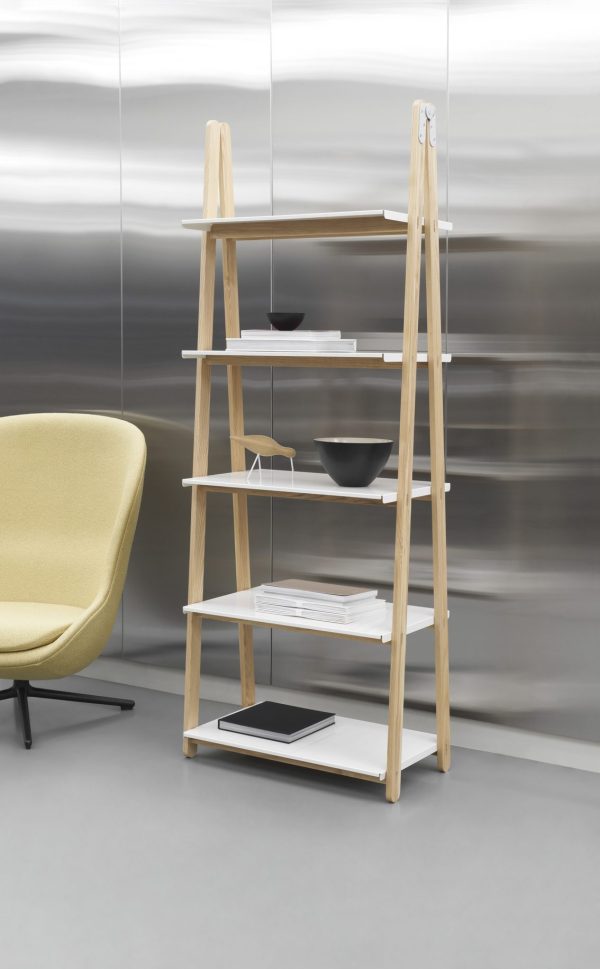 EN NORMANN COPENHAGEN - One Step Up Bogreol med fem hylder, med bøger og pynteting, ved siden af en gul stol. baggrunden har en sløret metallisk væg, der antyder bevægelse eller et hurtigt forløb.