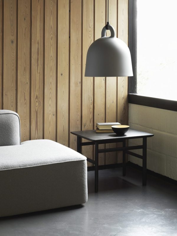 Et moderne hjørne af et værelse med en grå sofa, et sort sidebord med et par bøger og en NORMANN COPENHAGEN - Bell Lampe Ø55 hængende ovenover. rummet har træpanelvægge og mørkt gulv.