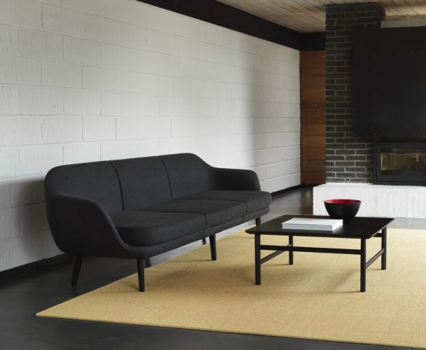 En moderne stue med en sort sofa, et firkantet sort sofabord med en rød skål, et stort beige tæppe og en pejs. Rummet viser et interiørdesign med hvidmalede murstensvægge og et mørkt træbjælkeloft.