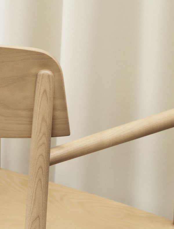 Nærbillede af en træstols ryg og armlæn, med let træåre synligt. Baggrunden byder på bløde beige gardiner og en cremefarvet væg. Stolen, der er stylet til TomorrowsDesign, er placeret indendørs med en varm, minimalistisk æstetik.