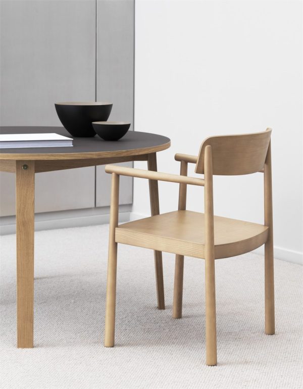 En spisestuestol i træ med armlæn placeret ved siden af et rundt bord med en sort skål på, i et rum med interiørdesignelementer som et lysegråt tæppe og hvide vægge.