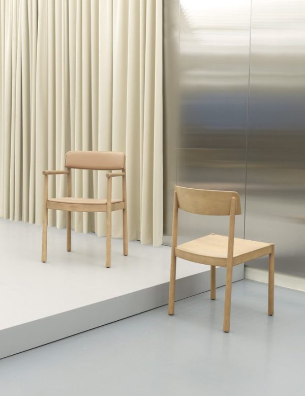 To minimalistiske møbler stole med træstel og beige sæder, den ene med armlæn, placeret på en grå platform mod en baggrund af bleggule gardiner og et metallisk panel.