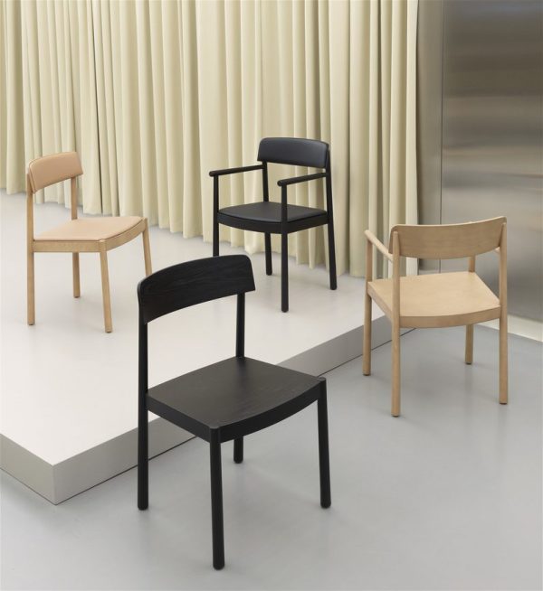 Fire minimalistiske møbler stole med forskellige designs placeret på et gråt gulv mod en kulisse af creme gardiner og en metallisk elevatordør.
