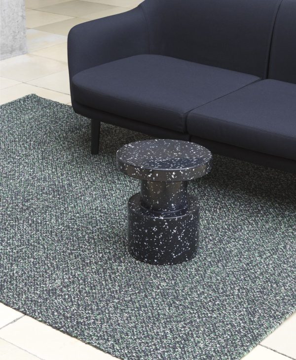 En moderne mørkfarvet sofa parret med et rundt, plettet sort sidebord på et tekstureret blågrønt og sort tæppe i minimalistiske indendørs omgivelser med NORMANN COPENHAGEN - Polli Gulvtæppe 100x200.