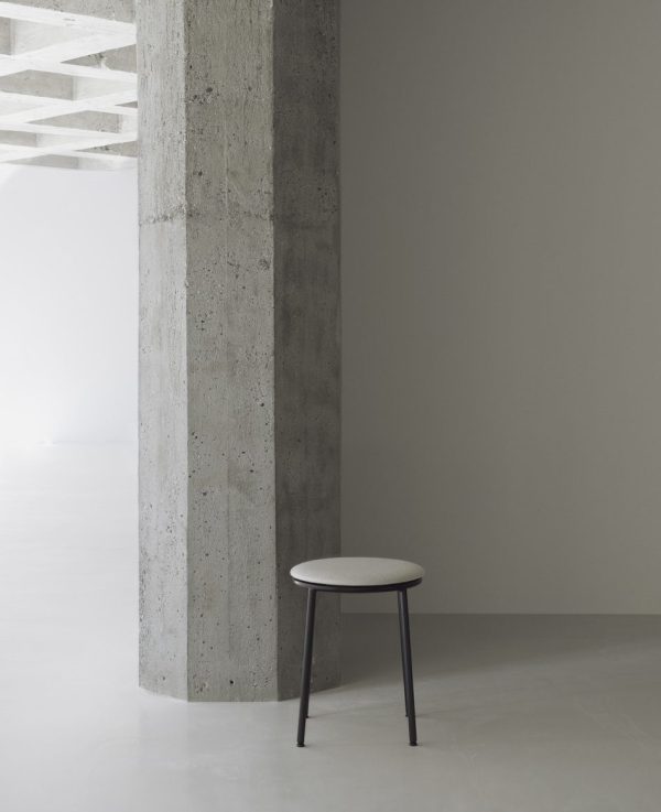 Et minimalistisk interiør med en grå betonsøjle med en lille, rund skammel placeret ved siden af. Gulvet er en glat, lysegrå overflade, og loftet har synlige bjælker.