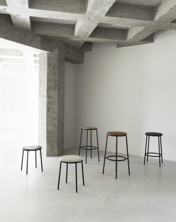 Et minimalistisk rum med et poleret betongulv, med synlige betonbjælker og fire forskellige designmøbler-skamler placeret op ad en hvid væg.