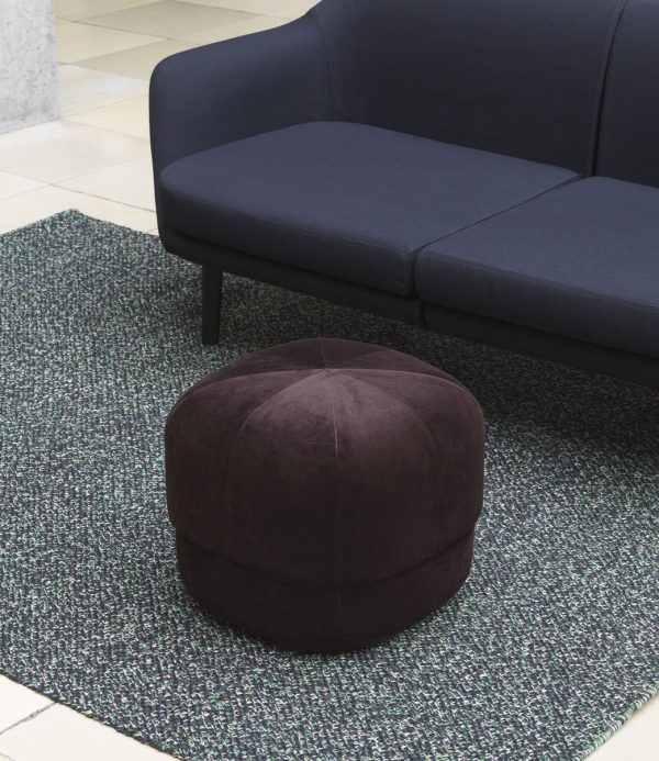 En mørkeblå sofa med en plys, mørkelilla rund skammel placeret på et NORMANN COPENHAGEN Polli Gulvtæppe 170x240 tæppe. Indstillingen antyder en moderne, minimalistisk stueindretning.