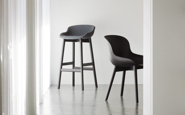 To NORMANN COPENHAGEN - Hyg Barstol 65 i et minimalistisk hvidt rum; den ene er en høj barstol og den anden en standardstol, begge i mørke farver, placeret nær et gennemsigtigt gardinvindue.