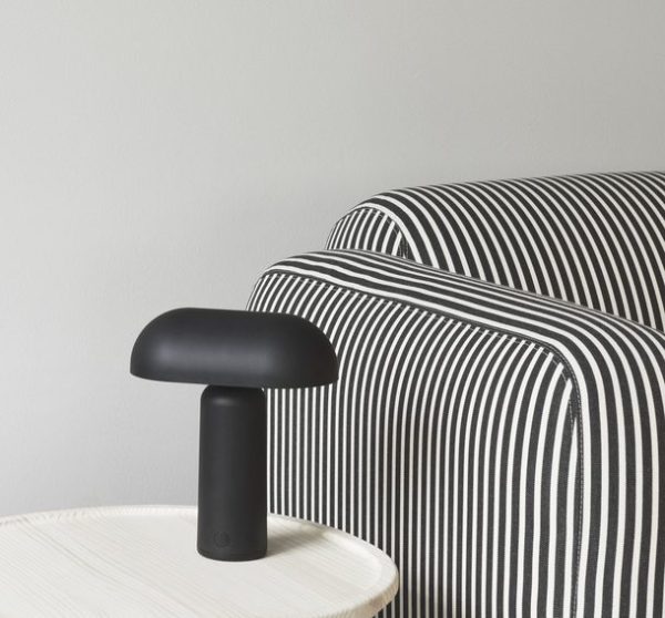 En moderne sort NORMANN COPENHAGEN - Porta Bordlampe sidder på et hvidt ribbet bord ved siden af en sofa med et fedt sort-hvidstribet mønster. baggrunden er en lysegrå farve, hvilket giver en neutral indstilling.