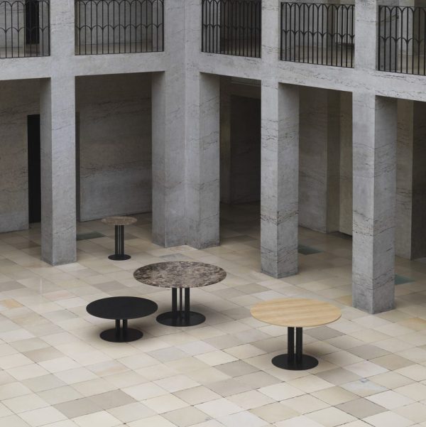 Et minimalistisk indendørs rum med et ternet gulv og tårnhøje stensøjler. Der er fire cirkulære borde med forskellige højder og finish, herunder træ og marmor, placeret centralt i dette møbler design.