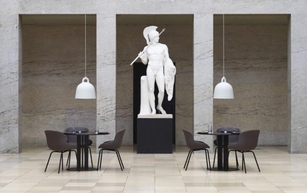 En marmorstatue af en nøgen mandsfigur står på en piedestal i en moderne lobby med tre buegange. To borde med designmøbler stole er placeret i nærheden, under hængende kuppellamper.