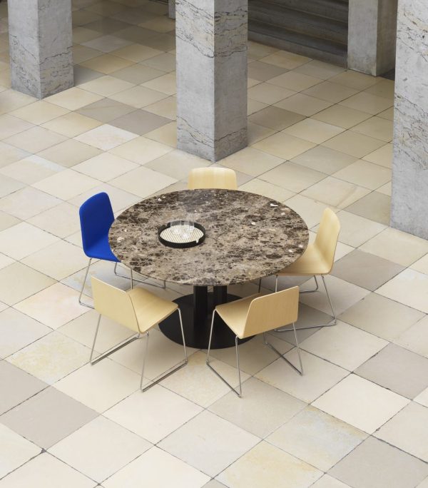 Et moderne cirkulært bord med granitplade og sort bund, omgivet af fire NORMANN COPENHAGEN - Just Stol stole i blå og guld, sat i et rummeligt område med klinkegulv og stensøjler.