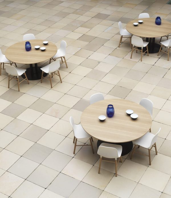 Et minimalistisk cafeteria med store runde træborde og hvide designmøbler stole. Hvert bord har en blå glasvase og hvide tallerkener. Gulvet er flisebelagt i beige og cremet ternet mønster.