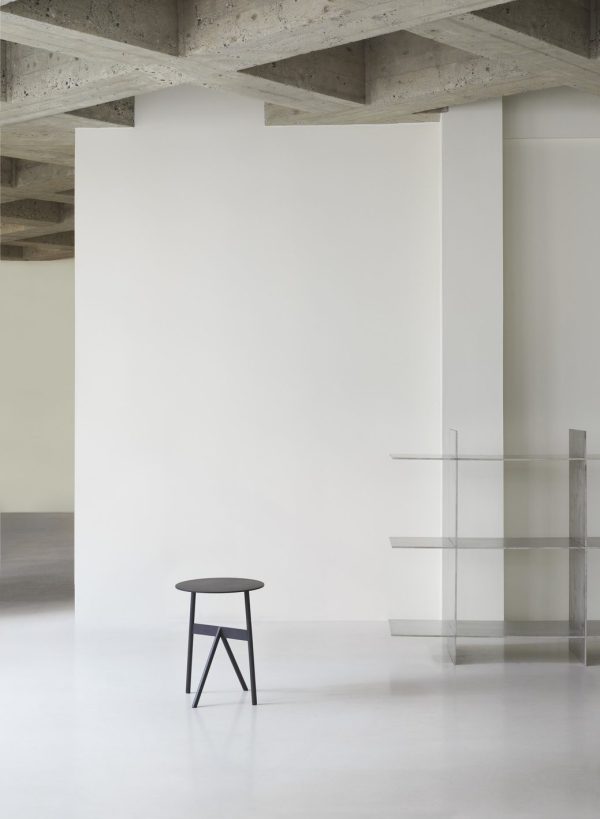 Et minimalistisk interiør med en NORMANN COPENHAGEN - Stock Bord Ø37 centreret foran en stor, blank hvid væg, med synlige grå betonbjælker over og en delvis udsigt til en glashylde til højre.