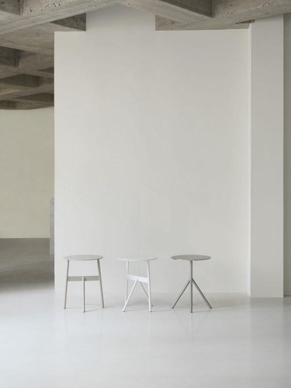 Et minimalistisk interiør med to små taburetter og en NORMANN COPENHAGEN - Stock Bord Ø37 nær en stor, blank hvid væg i et rum med betonloft. rummet er tomt og lyst, hvilket understreger enkelhed og moderne design.