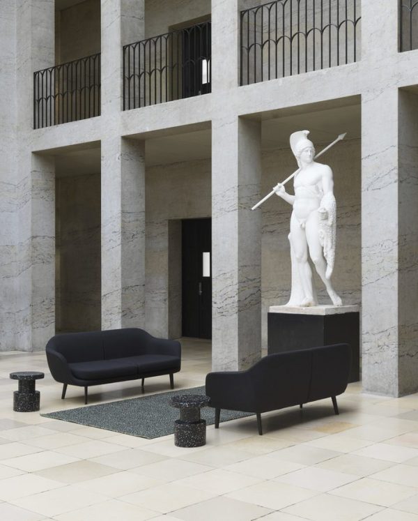 En hvid marmorstatue af en kriger, der holder et spyd og skjold, står i en minimalistisk lobby med to mørke sofaer og sorte cylindriske sideborde på et NORMANN COPENHAGEN - Polli Gulvtæppe 100x200 tæppe. baggrunden har en arkade i flere niveauer med balkoner.