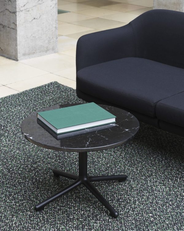 En moderne sort sofa ved siden af et lille rundt marmorbord med en grøn hardcover bog på toppen, placeret på et tekstureret grønt tæppe med en TomorrowsDesign-søjle i baggrunden.