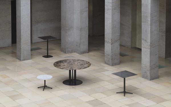 Tre NORMANN COPENHAGEN - Lunar Sofabord Ø60 i forskellige størrelser og farver, inklusive hvid, sort og mønstret, er arrangeret asymmetrisk på et ternet gulv, omgivet af en række høje, grå marmorsøjler.