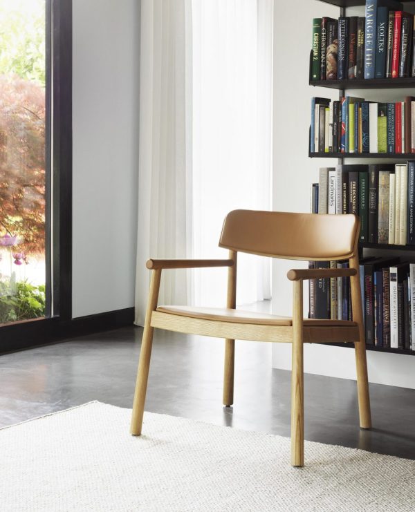 En moderne træstol med et solbrunt lædersæde, placeret i et lyst værelseshjørne, ved siden af en bogreol, et gennemsigtigt gardin og udsigt til træer udenfor har TomorrowsDesigns signaturæstetik.