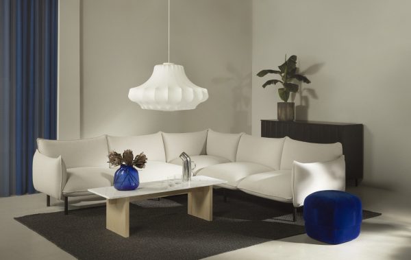 En stilfuld moderne stue med en ekspansiv NORMANN COPENHAGEN - Ark Modulsofa 100 Venstre, et firkantet sofabord i træ, en fed blå skammel og en stor pendel. En credenza i mørkt træ og gardiner komplementerer rummets neutrale palet.