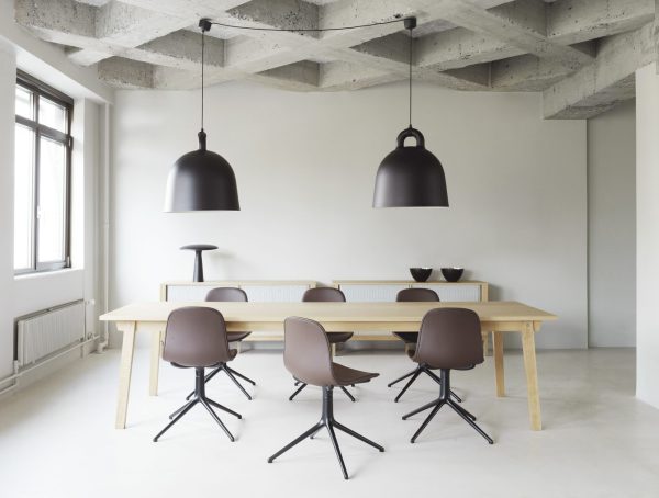 Minimalistisk spisestue med lyst træbord, brune stole, store sorte pendellamper og synligt betonloft. naturligt lys strømmer ind fra et vindue til venstre og fremhæver NORMANN COPENHAGEN - Bell Lampe Ø55, der hænger over bordet.