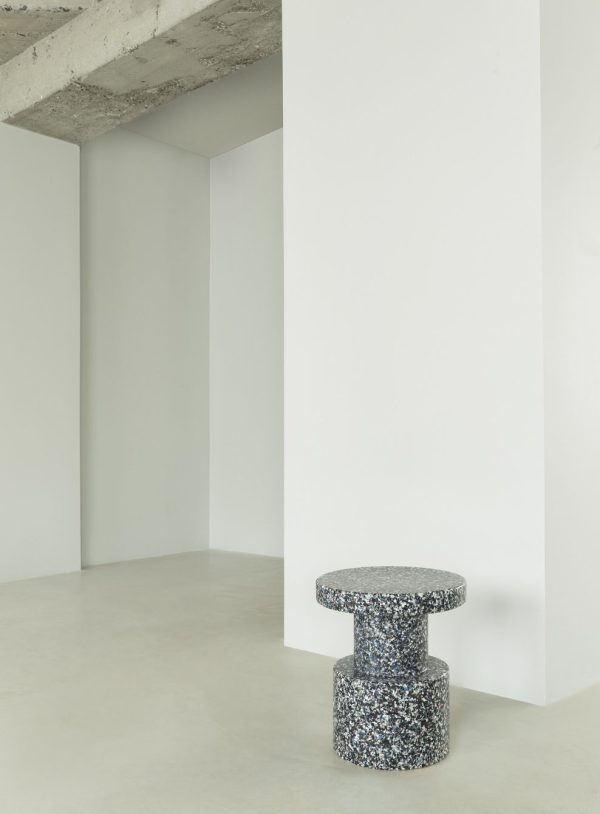 Et minimalistisk moderne rum med en hvid væg og søjle på et gråt betongulv, med en NORMANN COPENHAGEN - Bit Skammel på højre side. skamlen har en cylindrisk bund og en rund top.