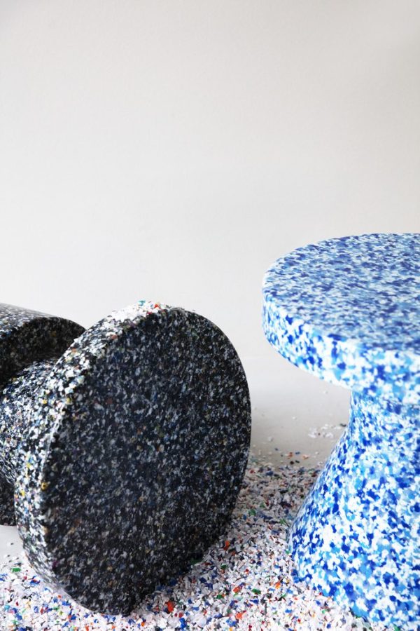To NORMANN COPENHAGEN - Bit Skammel Cone taburetter, den ene i sort med hvide pletter og den anden i blå med flerfarvede pletter, stående på et subtilt terrazzogulv i et rum med almindelige hvide vægge.