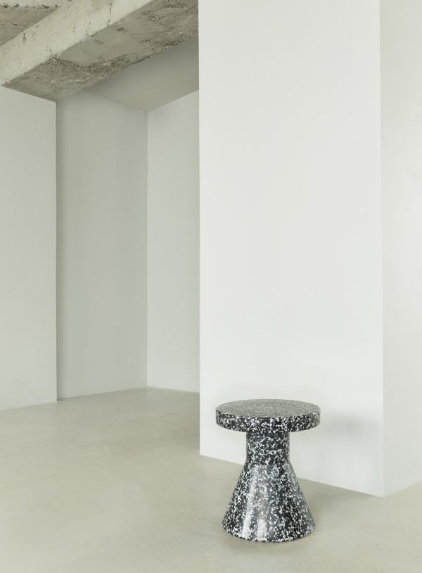 Et minimalistisk interiør med en NORMANN COPENHAGEN - Bit Skammel-kegle sat mod en almindelig hvid væg og et gråt betongulv, med en del af loftet synligt.