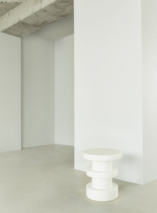 Et minimalistisk indvendigt rum med en hvid NORMANN COPENHAGEN - Bit Skammel Stack med teksturerede overflader, sat mod den vinkelgeometri af hvide vægge og et gråt betonloft.