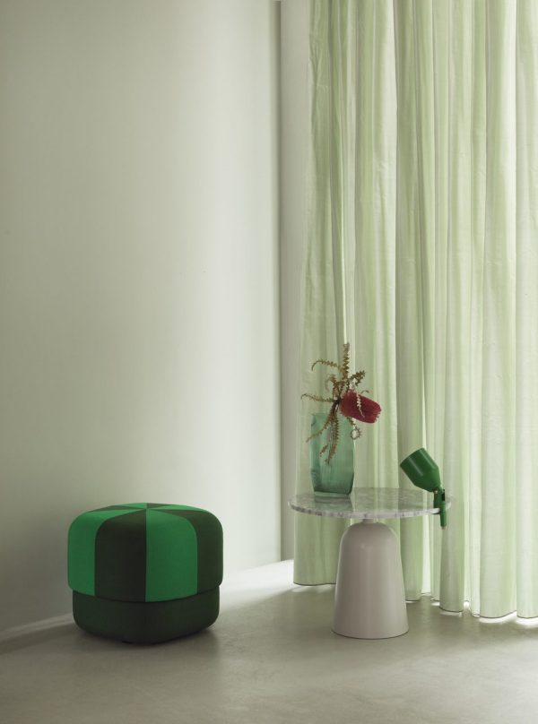 Et minimalistisk interiør med en NORMANN COPENHAGEN - Circus Puf Duo Lille, et sidebord i glas med en vase med blomster og en høj hvid lampe, alt sammen sat mod et blødt grønt gardin og en hvid væg.