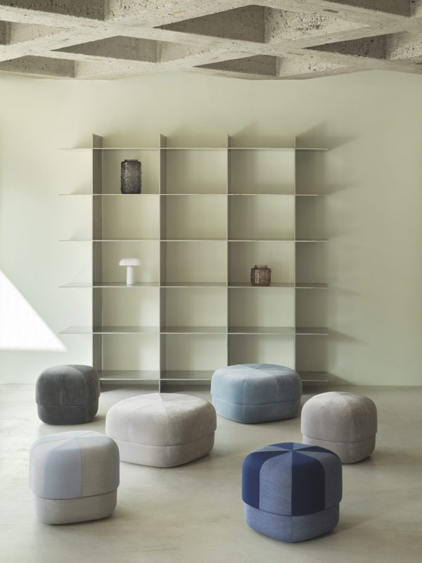 Et minimalistisk interiør med en stor, tom hvid reol mod en betonvæg, med seks NORMANN COPENHAGEN - Circus Puf Duo Lille i nuancer af grå, blå og lavendel arrangeret foran.