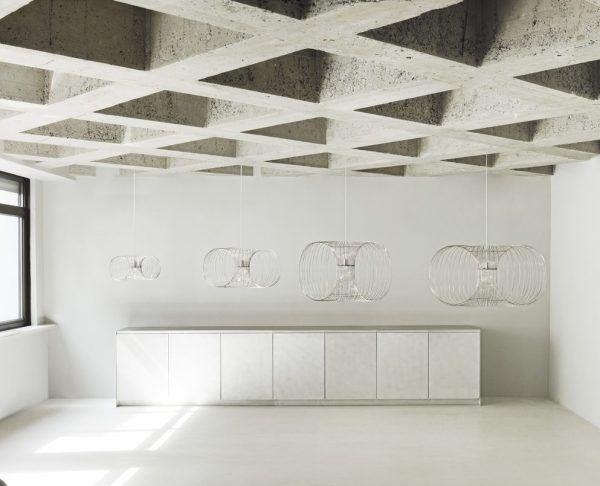 Et minimalistisk moderne køkken med hvide skabe, lang bordplade og NORMANN COPENHAGEN - Coil Lampe Ø76 pendellamper. fritlagte betonbjælker i loftet og et vindue, der slipper naturligt lys ind.