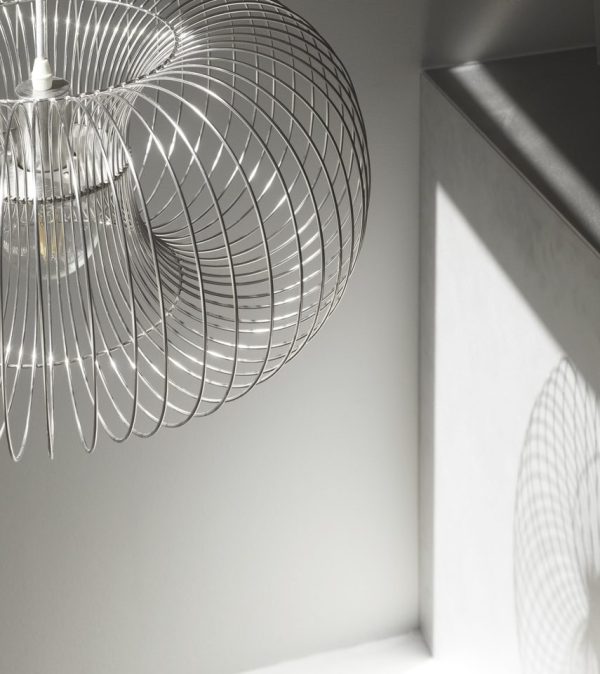 En moderne pendel med et spiraltrådsdesign hænger i et rum og kaster indviklede skygger på den hvide væg. Det omgivende lys skaber en blød, minimalistisk interiør-æstetik.
