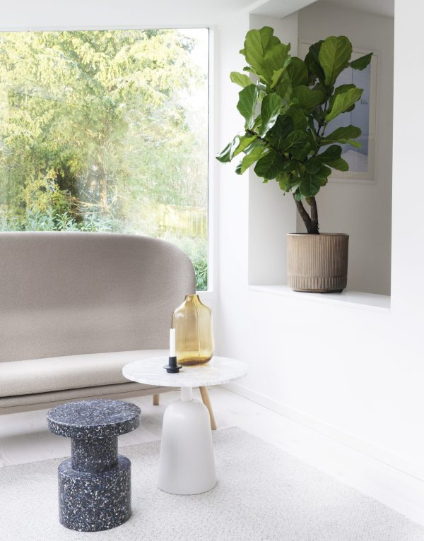 Et moderne stuehjørne med en beige sofa, et rundt hvidt bord med en gul vase, NORMANN COPENHAGEN - Bit Skammel skamler og et stort figentræ i potte ved et vindue med udsigt over en grøn have.