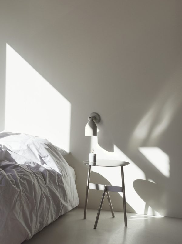 Et minimalistisk soveværelse med blødt morgenlys, der kaster skygger på en hvid væg. et lille sidebord rummer en NORMANN COPENHAGEN - Klip Væglampe og et krus ved siden af en uopredt seng med en krøllet hvid dyne.