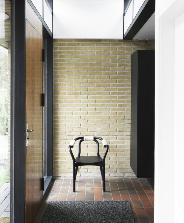 En moderne gang med terracotta gulvfliser og en gul murstensvæg. en enkelt NORMANN COPENHAGEN - Knot Spisebordsstol er centreret mod væggen, under en trappe med metalrækværk. store glasdøre åbner udad til venstre.