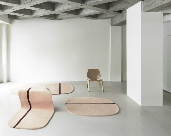 Moderne minimalistisk interiør med en enkelt træstol og tre NORMANN COPENHAGEN - Oona Gulvtæppe 140x140 tæpper med brune kanter på et gråt betongulv, under en synlig loftstruktur.