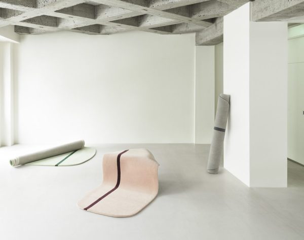 Et minimalistisk interiør med synligt betonloft og søjler. tre NORMANN COPENHAGEN - Oona Gulvtæppe 90x200 tæpper i forskelligt design lå på det glatte, lysegrå gulv.