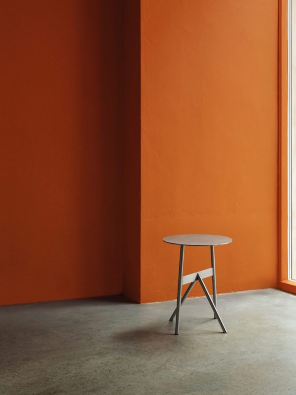 Et minimalistisk interiør med et NORMANN COPENHAGEN - Stock Bord Ø37 bord mod en levende orange væg, nær et vindue med sollys, der kaster lyse højdepunkter og skygger.